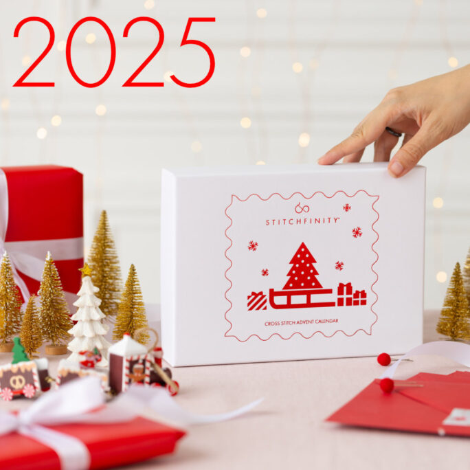 Image of the packaging of the 2025 STITCHFINITY cross stitch advent calendar showing the box, a hand reaching to pick it up and Christmas decorations